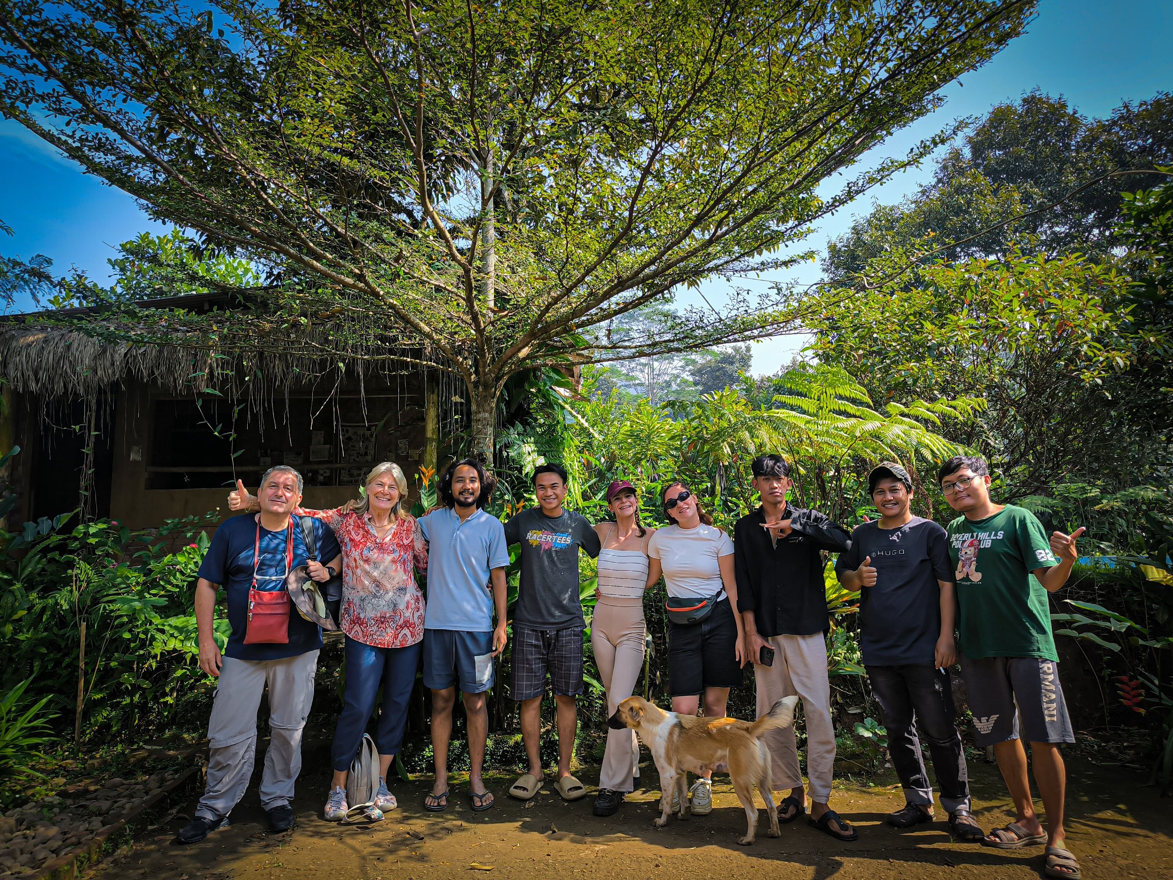 Un grupo de personas de pie al aire libre, con un árbol grande y abundante vegetación de fondo.
