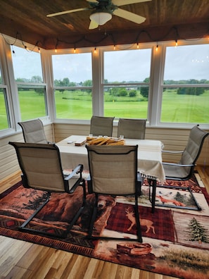 A dining area with a table, chairs, and a view of a green landscape through large windows.