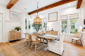 Un coin repas avec une table en bois, des chaises en osier, un luminaire suspendu et de grandes fenêtres ornées de rideaux blancs.