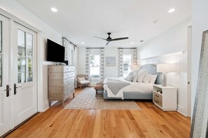 Une chambre à coucher comprenant un lit, un téléviseur, un ventilateur de plafond, une commode, une chaise et une fenêtre avec des rideaux.