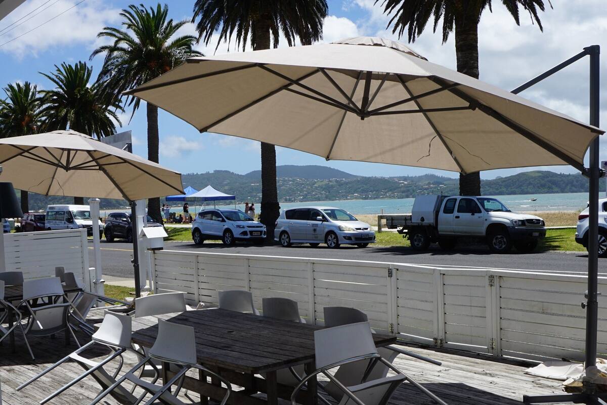 Un área de estar al aire libre con mesas y sillas bajo sombrillas, con vista a una playa con palmeras y coches estacionados.