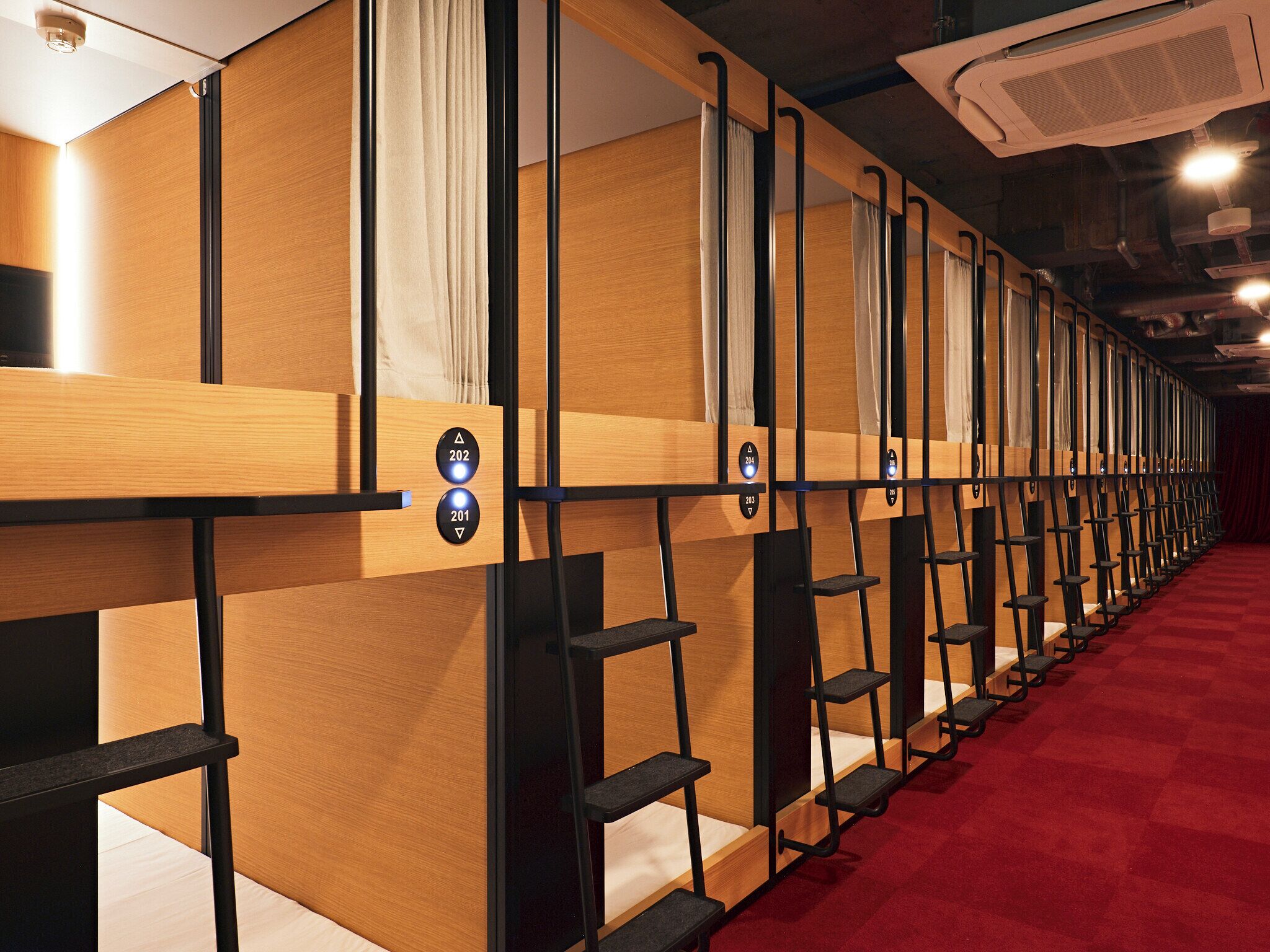 Reihe von Holzbunkbetten mit separaten Bedienfeldern, ein roten Teppichboden und eine Decke mit freiliegenden Balken und Leuchten.