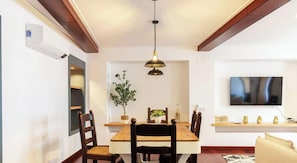A dining area with a wooden table, chairs, a hanging light fixture, a potted plant, and a mounted TV.