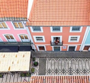 Un immeuble à plusieurs étages, doté d’un toit recouvert de tuiles rouges, d’un balcon où se trouve une personne, et d’un trottoir à motifs.