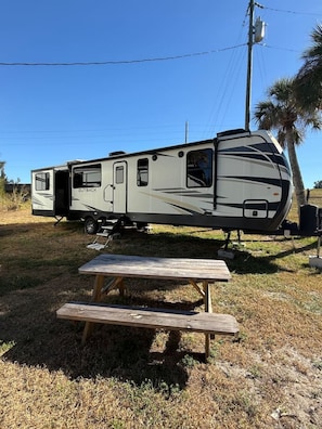 A recreational vehicle with a slide-out awning, a picnic table, and a palm tree in the background.