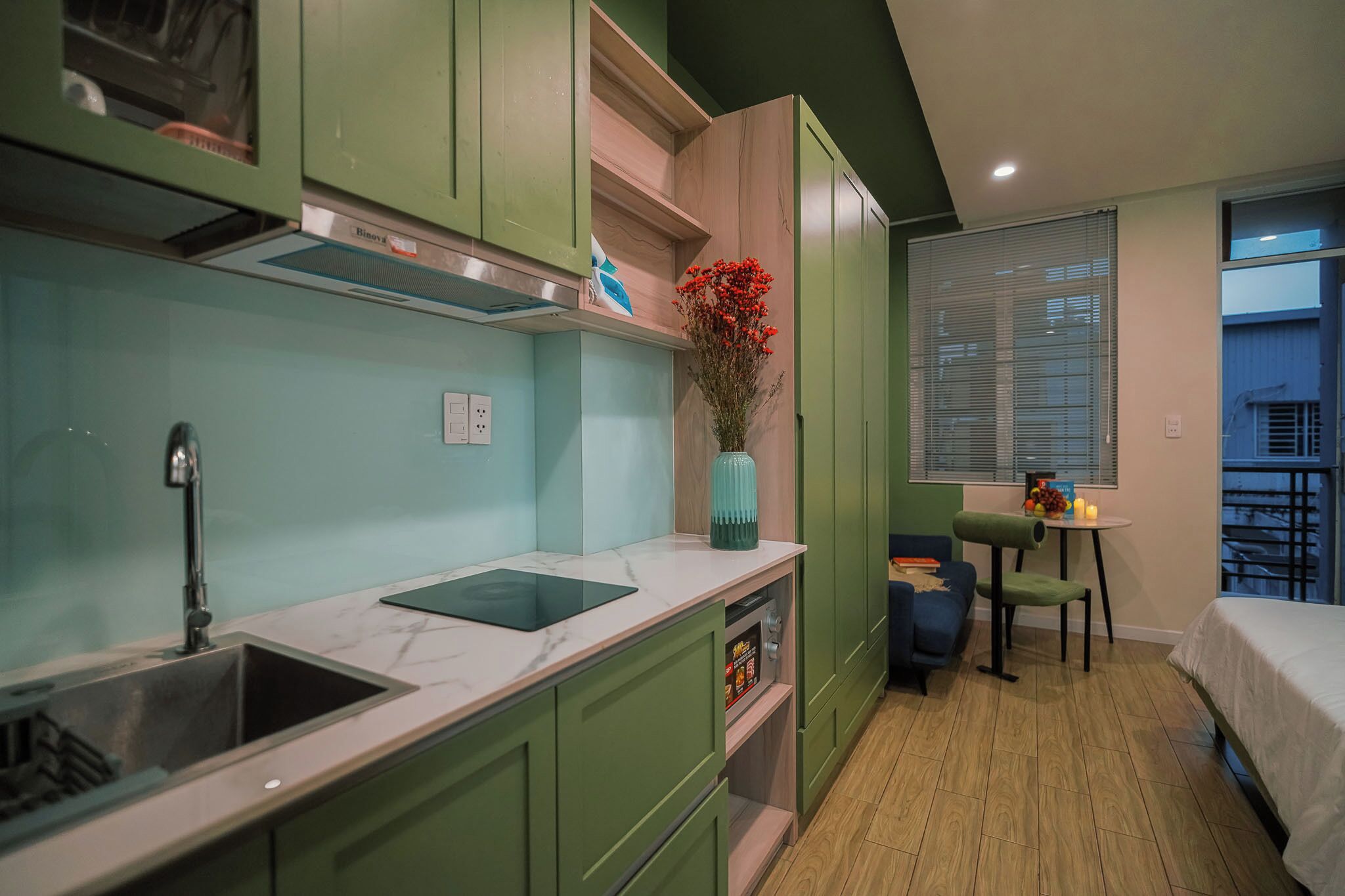 A compact kitchen with green cabinets, a marble countertop, a sink, and a vase with red flowers.