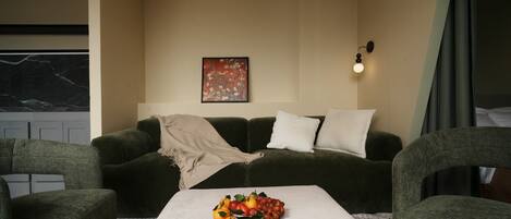 Een woonkamer met een bank, een bijzettafeltje met fruit, een schilderij en een wandlamp.