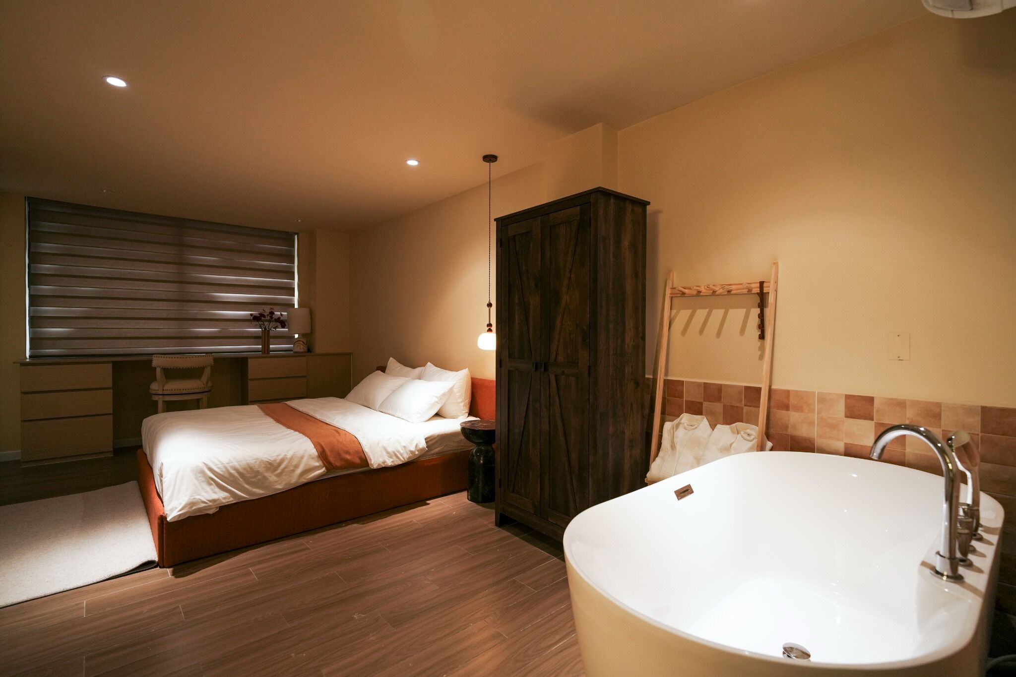 A bedroom with a bed, a wooden wardrobe, a bathroom with a bathtub, and a window with blinds.