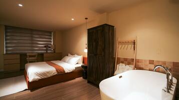 A bedroom with a bed, a wooden wardrobe, a bathroom with a bathtub, and a window with blinds.
