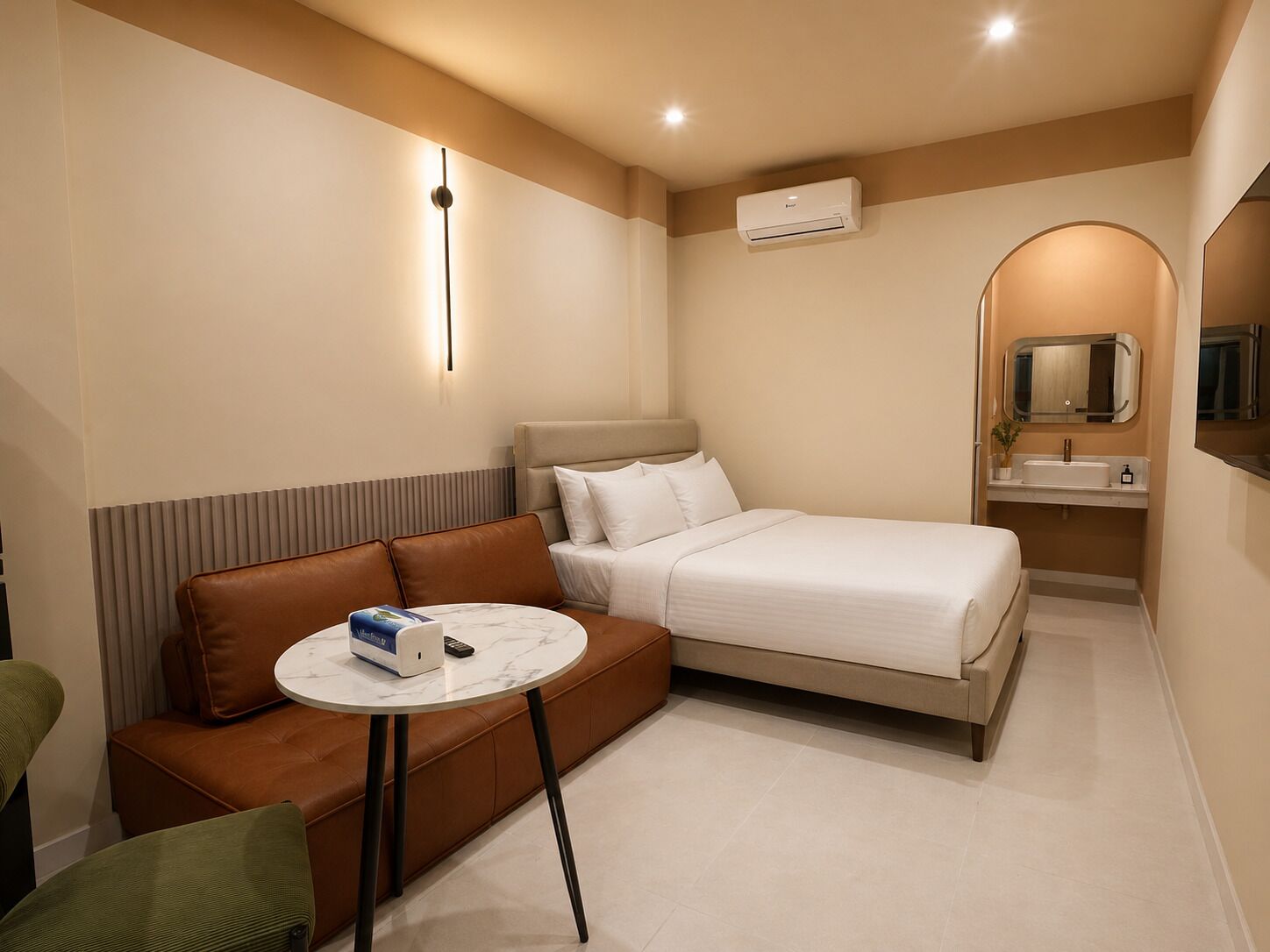 A hotel room with a bed, a sofa, a small table, a mirror, and an air conditioning unit.