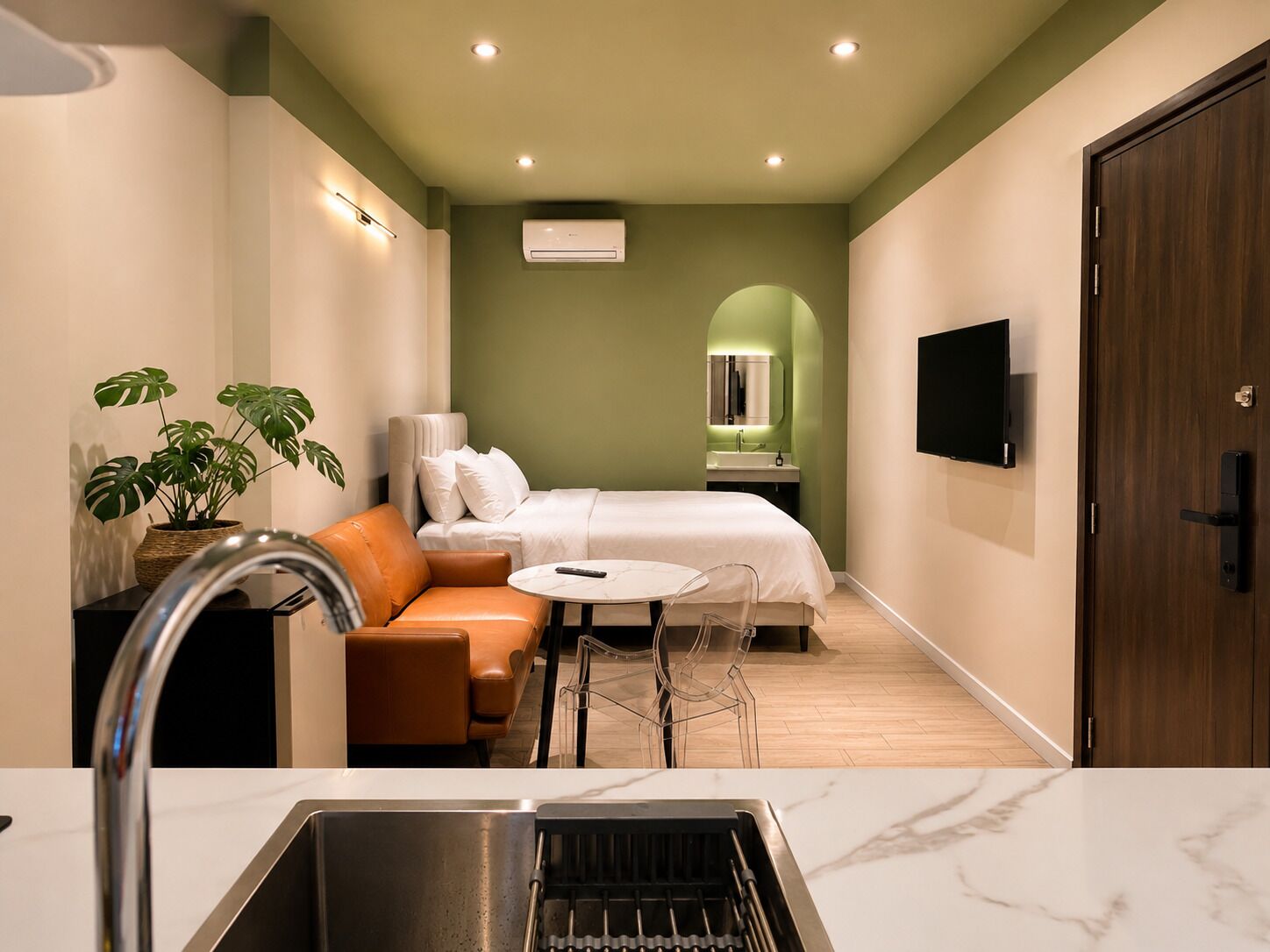Ein kleines Hotelzimmer mit Bett, einem Tischchen, einer Couch, einem Fernseher, einem Waschbecken und einer Topfpflanze.