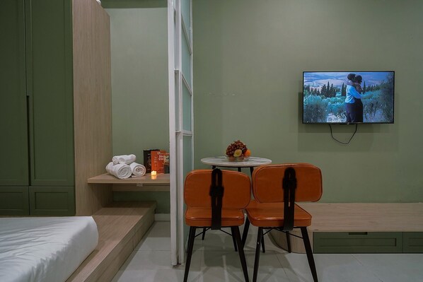 A hotel room with a bed, a TV on the wall, a small table with fruit, two orange chairs, and a shelf with towels and a candle. - PIN Nguyen Cu Trinh (Ho Chi Minh City)