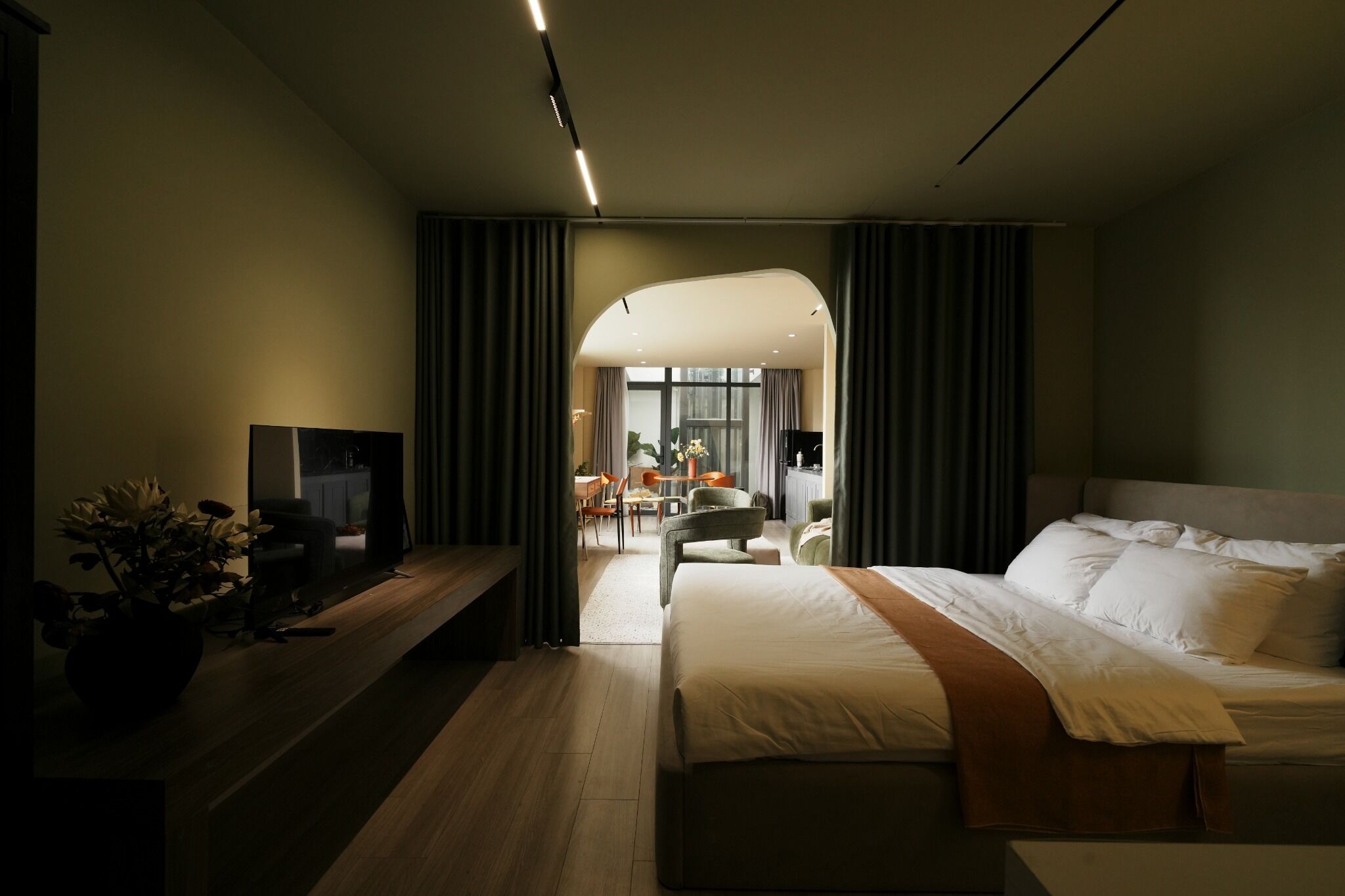 A modern hotel room with a large bed, a desk, and a view into the dining area.