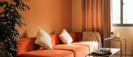 Una sala de estar acogedora con un sofá, una mesa de ajedrez, una planta en maceta y una ventana con cortinas.