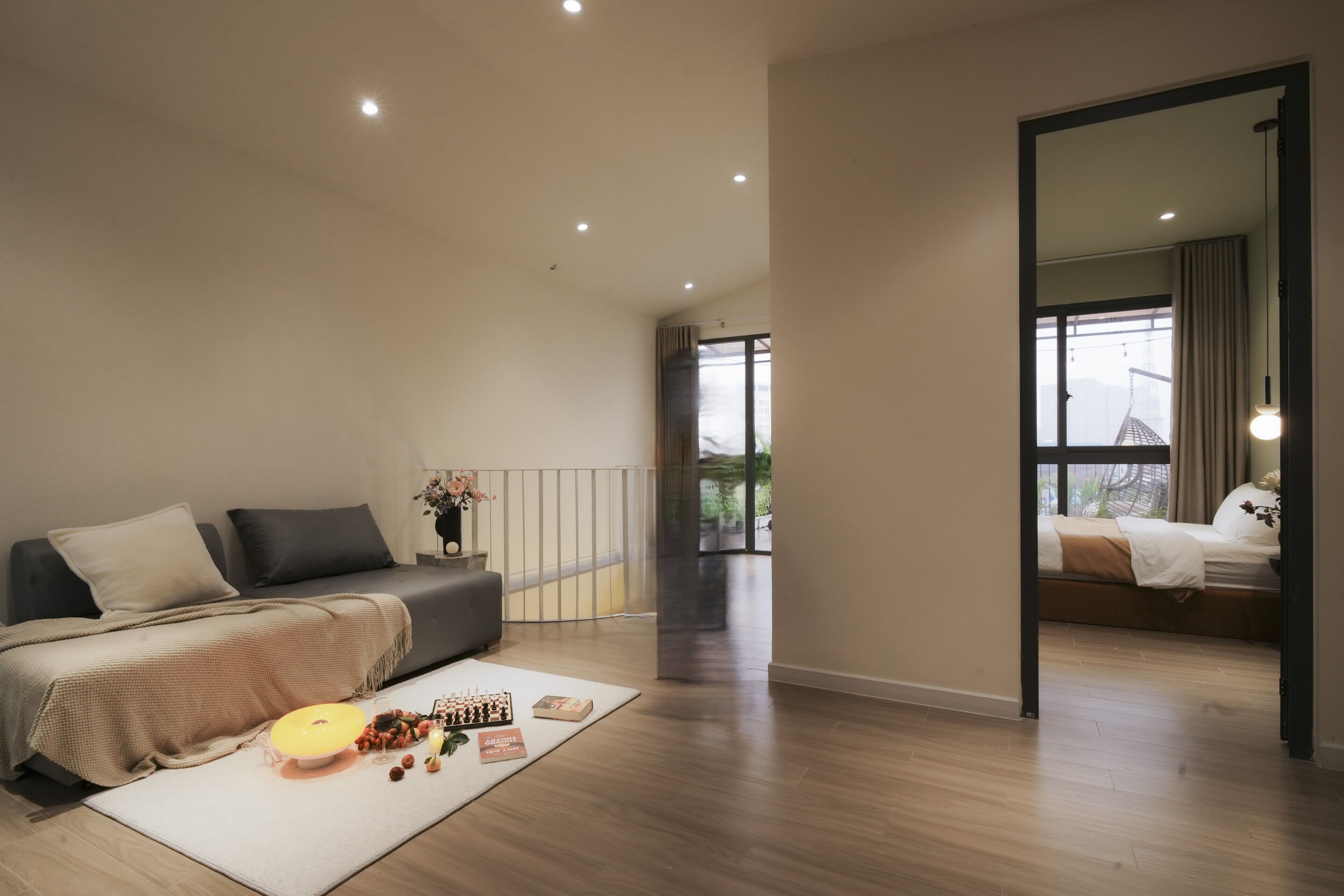 A modern bedroom with a sofa bed, a dining table with a bowl and books, and a large mirror reflecting a balcony.