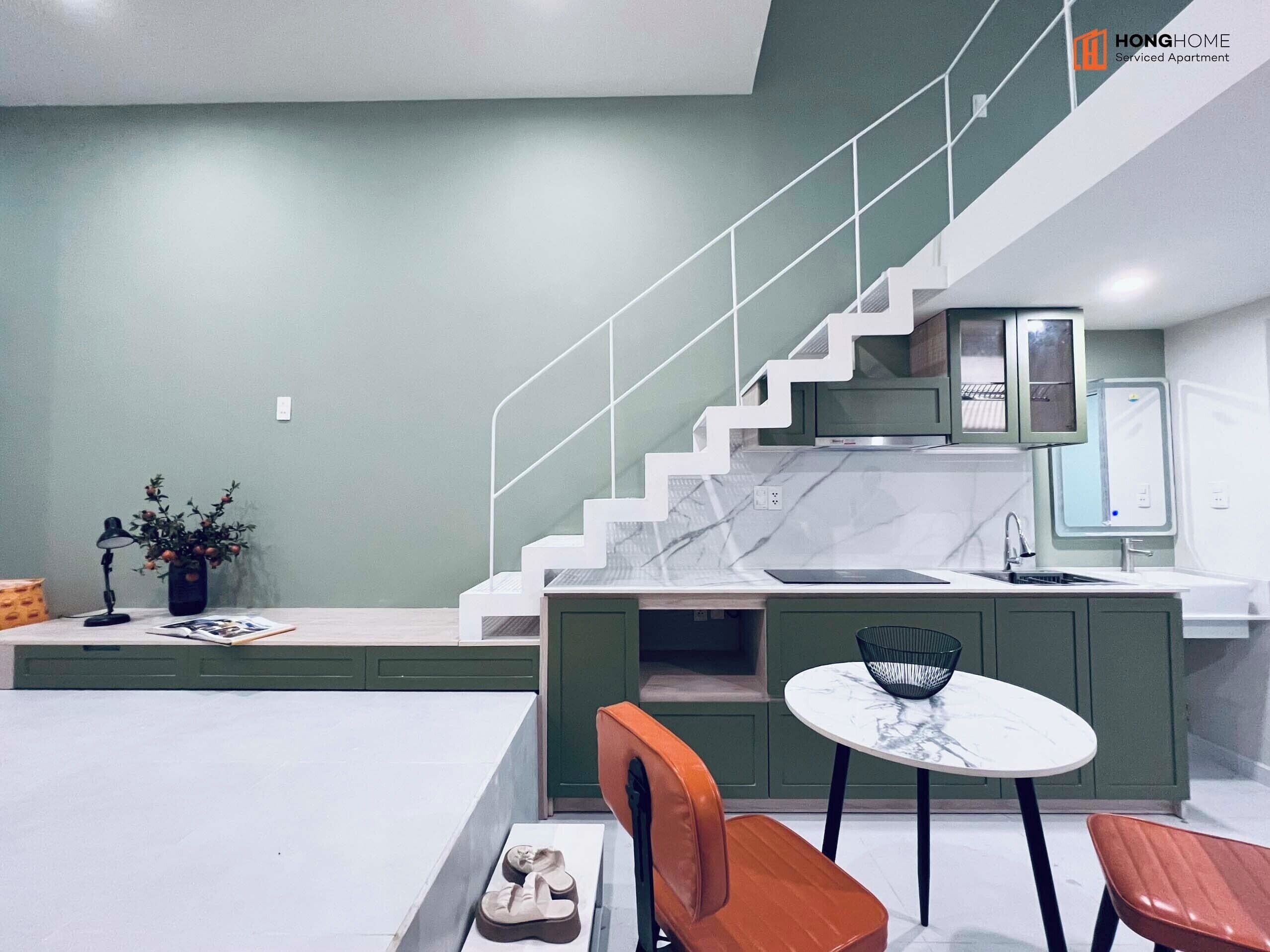 Una cocina moderna con encimera de mármol, armarios verdes y estufa integrada. Hay una zona de comedor con una mesa redonda y dos sillas.