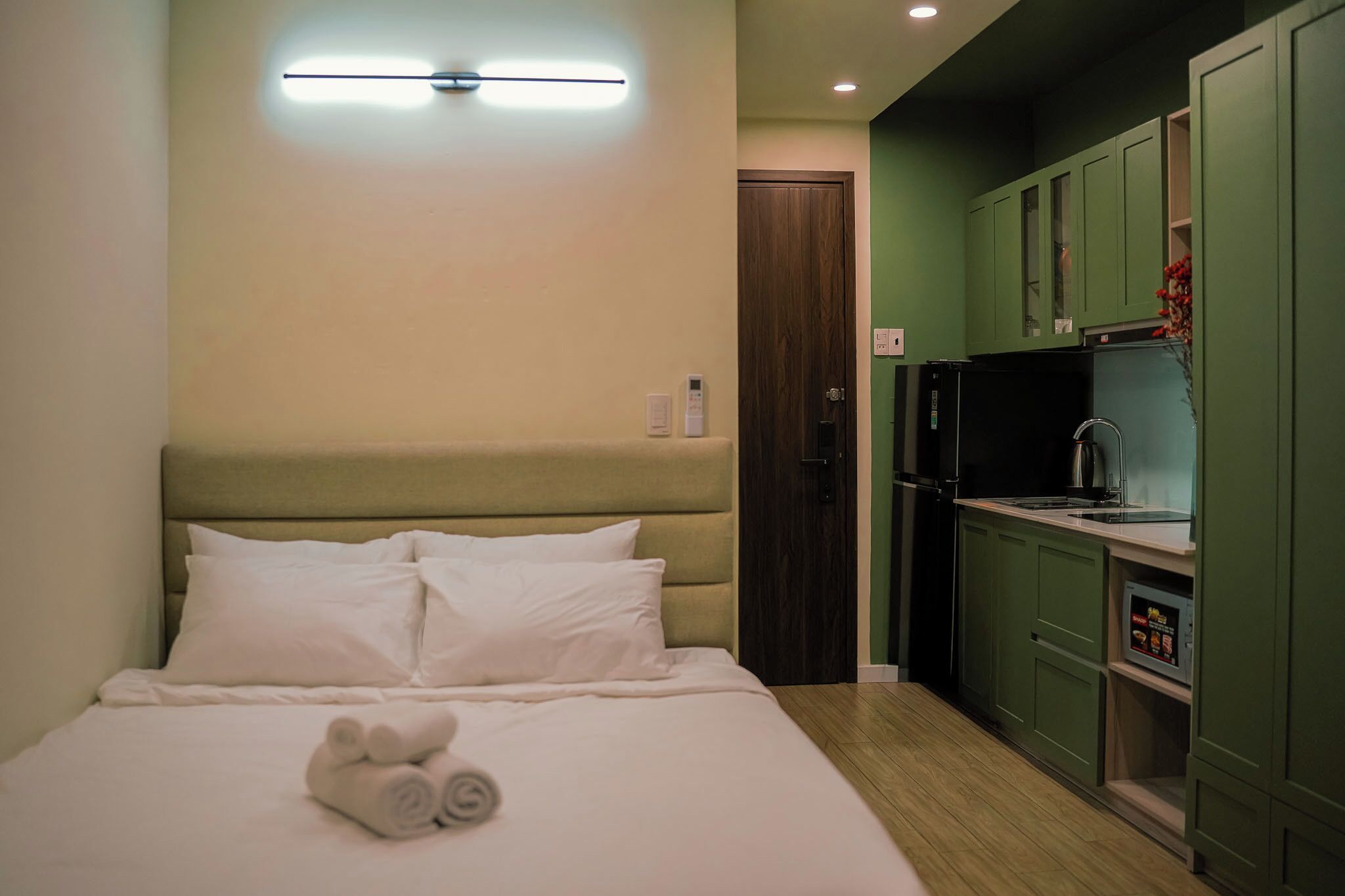 Een hotelkamer met een bed, een nachtkastje met een lamp, een kitchenette met een koelkast en een deur.