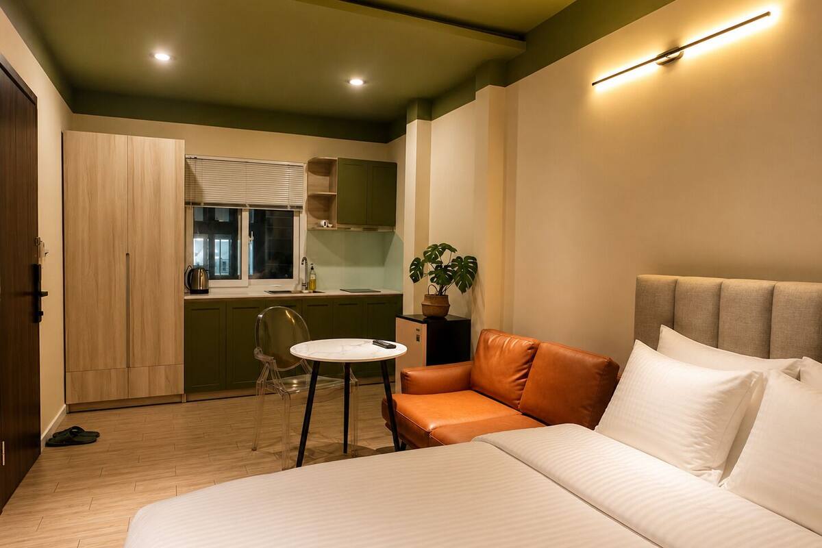 A bedroom with a bed, a small table, a sofa, a potted plant, and a kitchenette.