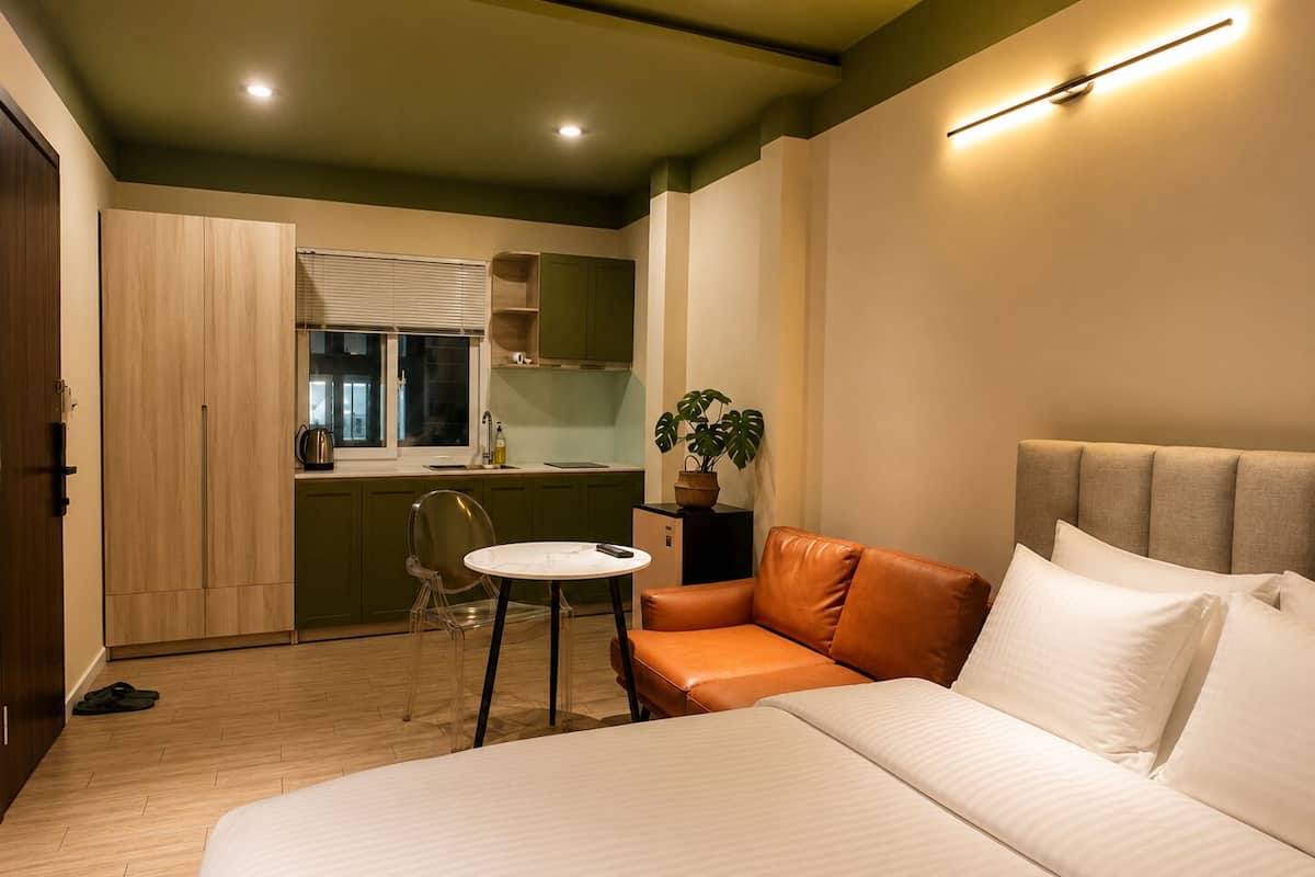 Een slaapkamer met een bed, een kleintje tafel, een bank, een plant in pot en een kitchenette.