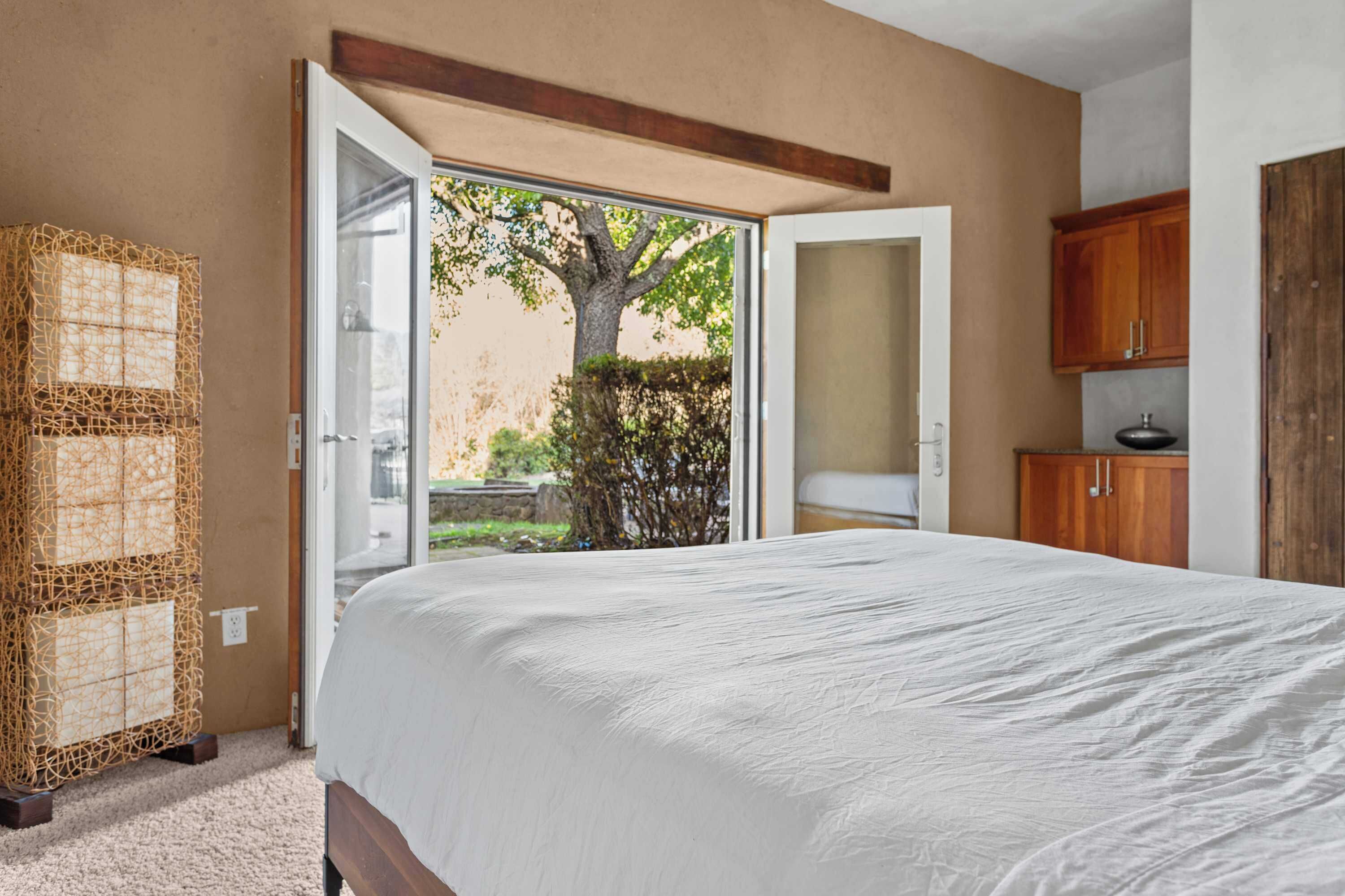 A bedroom with a large bed, an open door leading to a balcony, a wicker chair, and wooden cabinets.