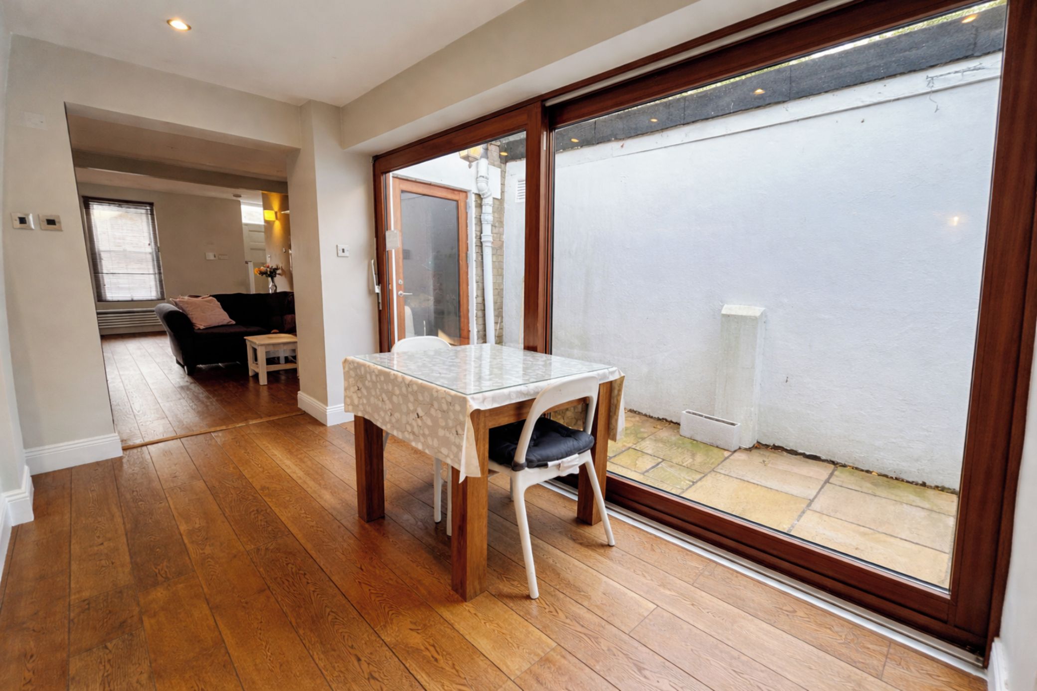 A room with a wooden floor, a table with a white tablecloth, a chair, and a sliding glass door leading to an outdoor area.
