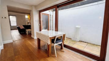A room with a wooden floor, a table with a white tablecloth, a chair, and a sliding glass door leading to an outdoor area.