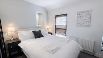 A neatly made bed with white linens, a black and white checkered pillow, and a folded white towel on the bed. There is a nightstand with a lamp and a small drawer. A window with blinds is visible, and a wall-mounted artwork is present.