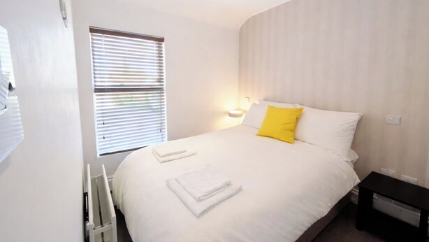 A hotel room with a bed, a bedside table, a lamp, and a window with blinds. - Contemporary 2BDR City Retreat (Dublin)