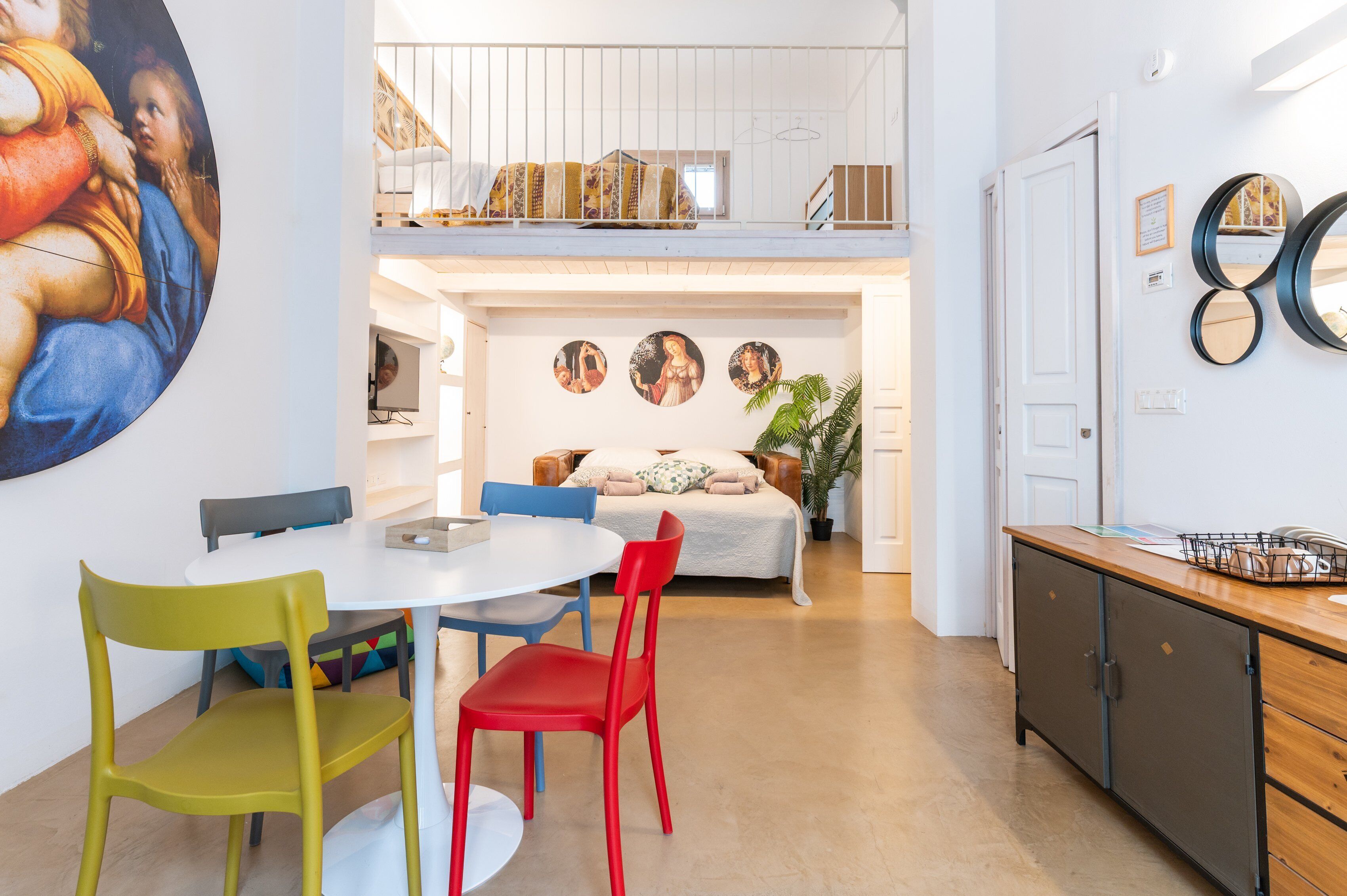 Un'area pranzo con un tavolo bianco e sedie colorate, un divano con una coperta decorata e una scala che conduce a un piano superiore.
