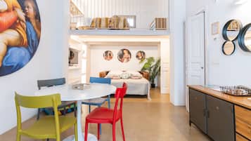 Un'area pranzo con un tavolo bianco e sedie colorate, un divano con una coperta decorata e una scala che conduce a un piano superiore.