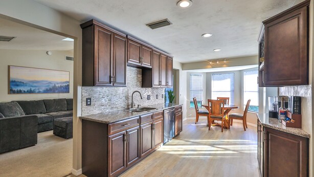 A modern kitchen with dark wood cabinets, a granite countertop, and a built-in island with a sink. There is a dining area with a table and chairs, and a living room with a couch and a painting on the wall. - Coastal Oasis! Hot Tub, Pool & Ping Pong Tables, Fenced Yard, Near Beach! (Port Orange)