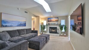 A living room with a gray sectional sofa, a fireplace, and a large painting of a forest. - Coastal Oasis! Hot Tub, Pool & Ping Pong Tables, Fenced Yard, Near Beach! (Port Orange)