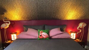 Una cama con una colcha gris texturizada, dos almohadas rosas, una almohada verde y dos luces de noche con forma de flamenco a cada lado.