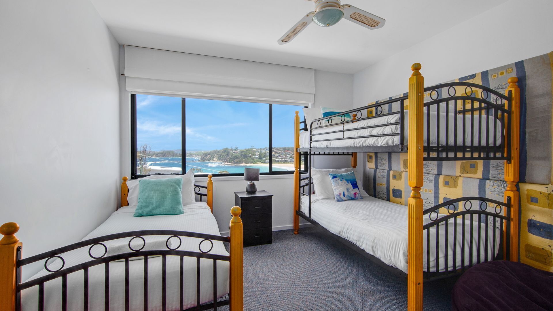 A room with a single bed, a bunk bed, a window with a view of the sea, and a ceiling fan.