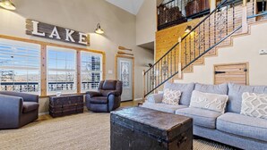 A spacious living room with a large window, a leather armchair, a grey sofa with patterned cushions, and a rustic wooden coffee table. - Emerald Point, Lakefront with Gorgeous Views! (McAlester)