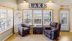A cozy living room with a fireplace, leather chairs, a wooden chest, and a large window with blinds. - Emerald Point, Lakefront with Gorgeous Views! (McAlester)