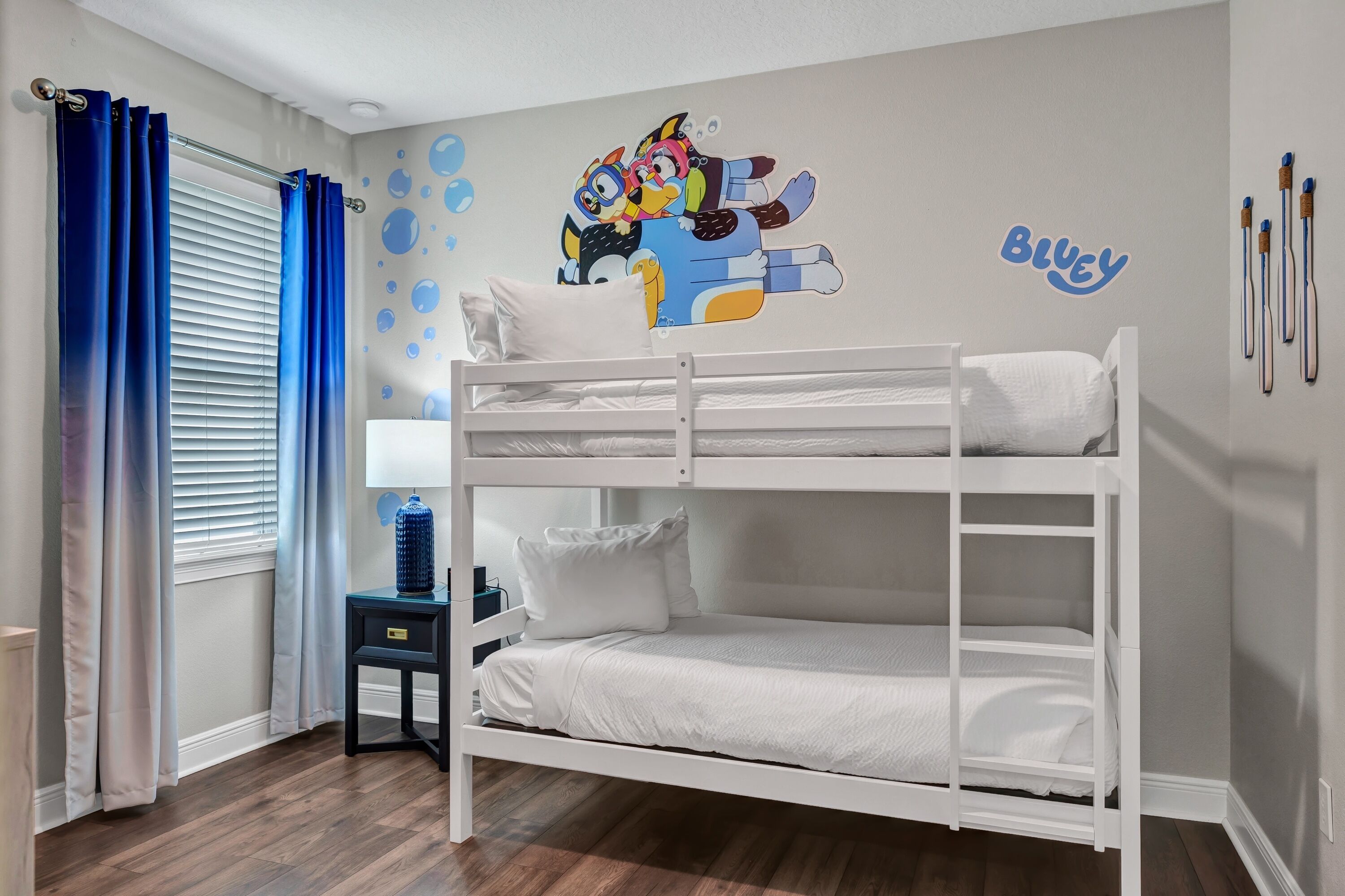 Een slaapkamer met stapelbed, een muurschildering, een raam met blauwe gordijnen en een klein tafeltje met een waterfles.