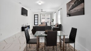 Een moderne woonkamer met een glazen eettafel, zwarte stoelen, een bank en een aan de muur bevestigde televisie.