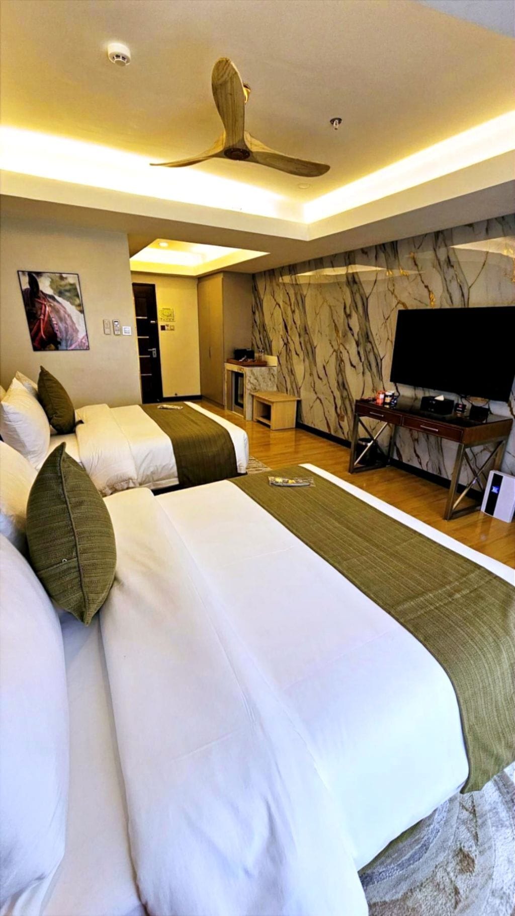 A hotel room with two beds, a desk, a television, and a ceiling fan.