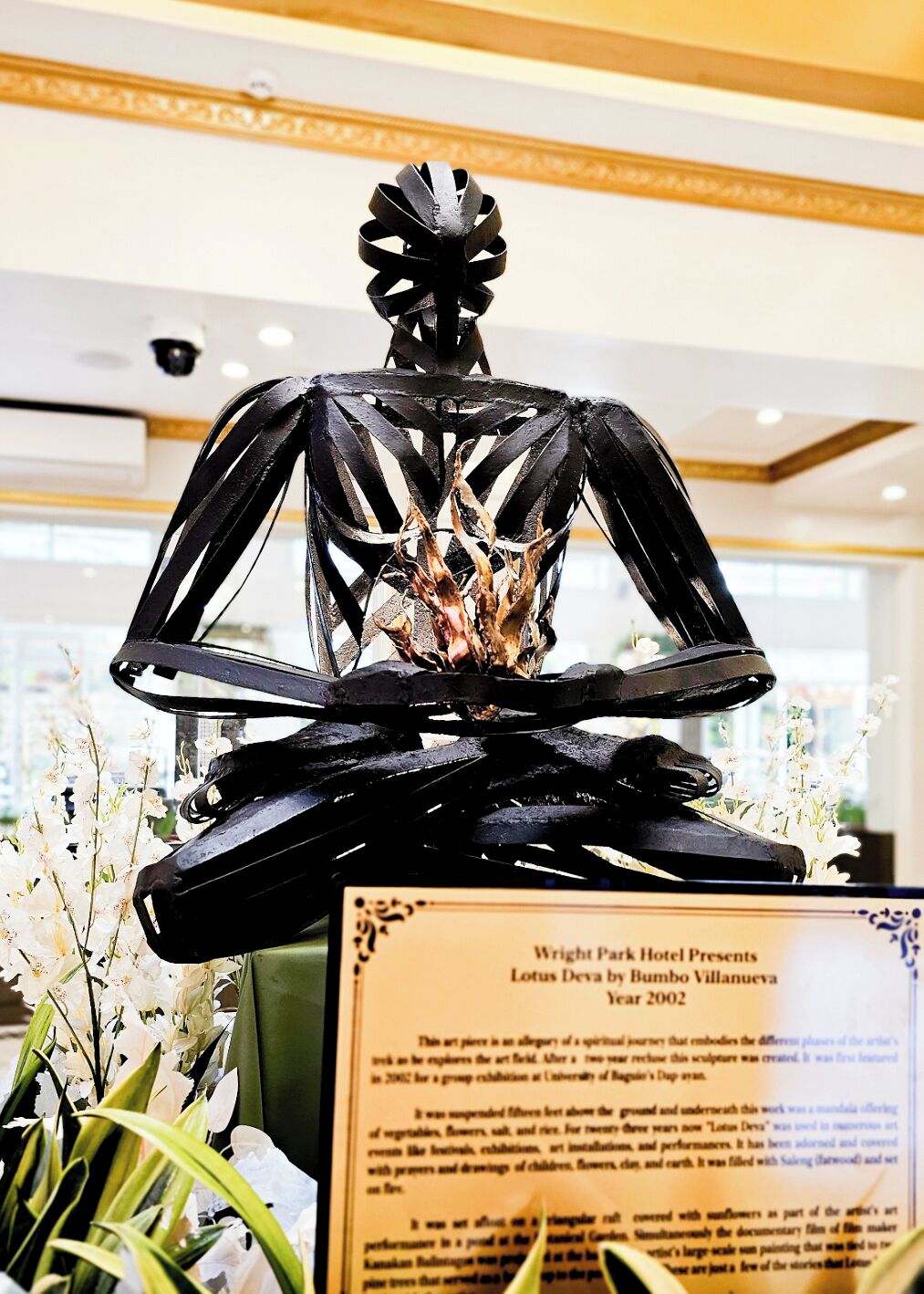A sculpture of a meditative figure with a lotus flower, displayed in a hotel lobby with a plaque describing it as 'Lotus Deva by Bumbo Villanueva, Year 2002'.