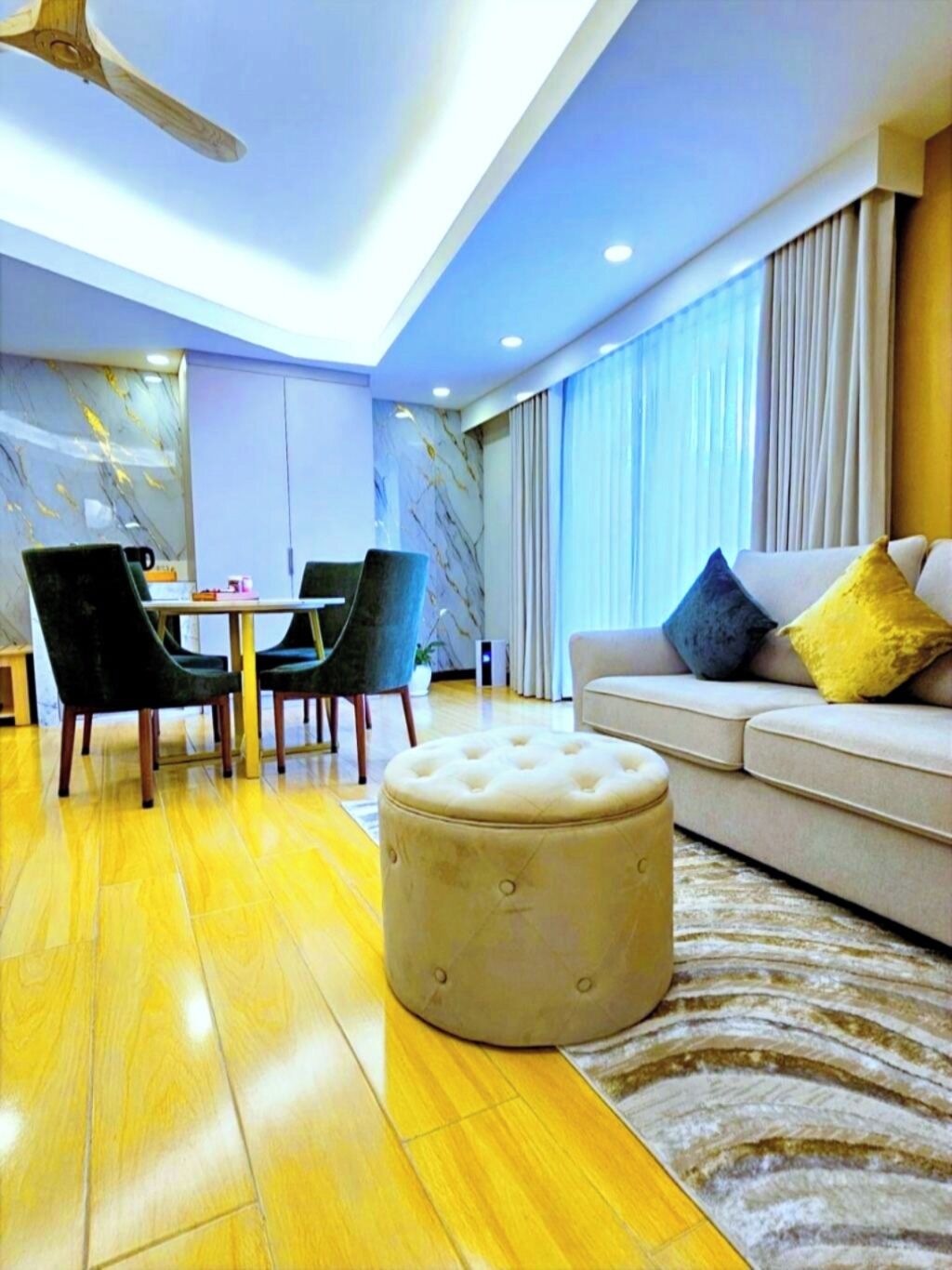 A modern living room with a wooden floor, a round ottoman, a sofa with cushions, and a dining area with chairs and a table.