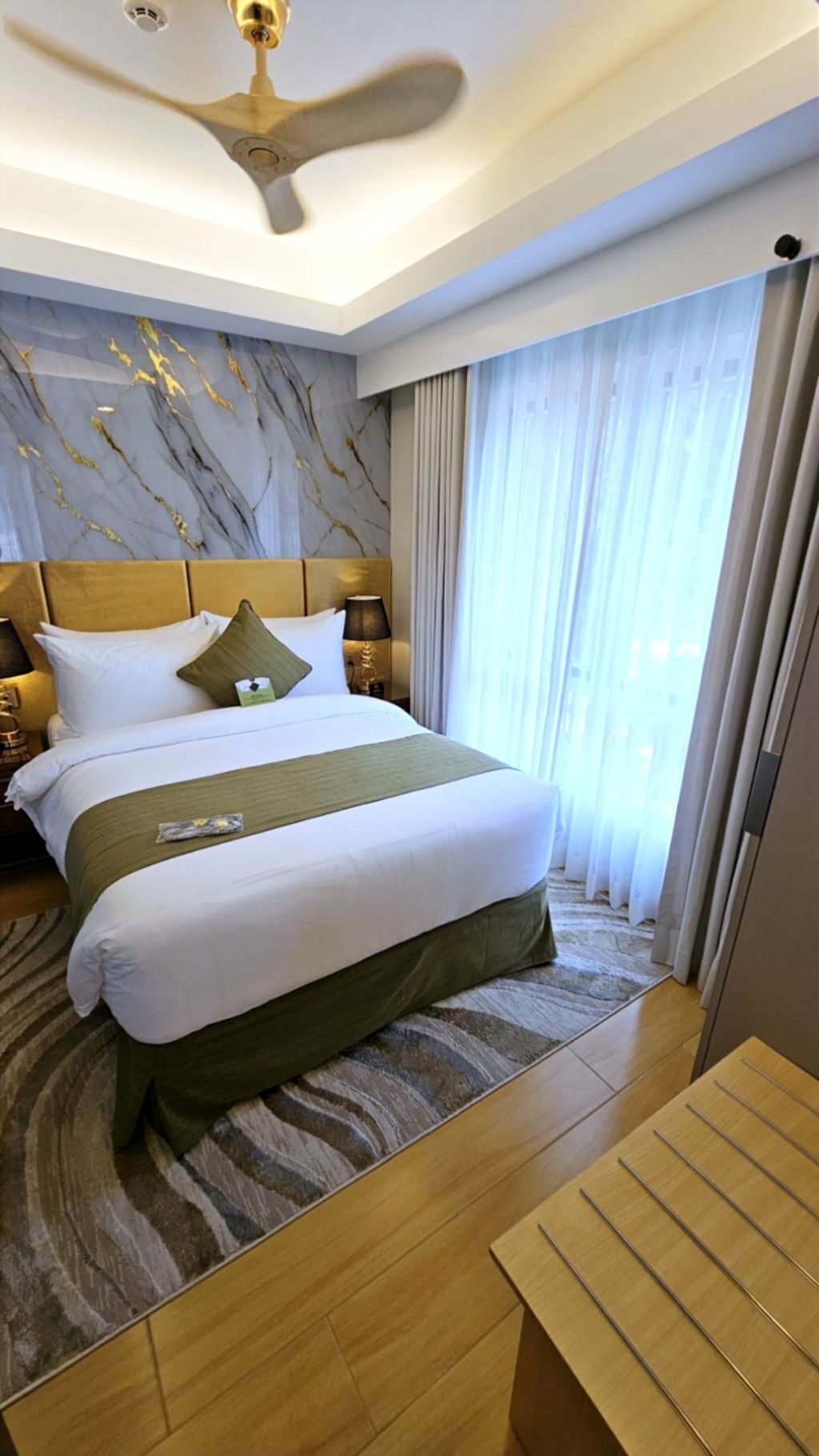 A hotel room with a large bed, a ceiling fan, a marble-patterned wall, and a view of the city through the window.