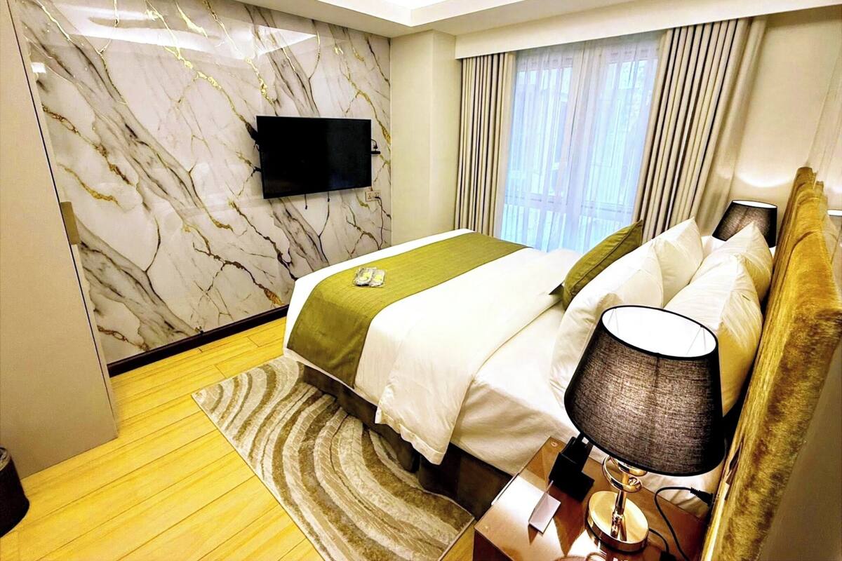 Ein Hotelzimmer mit Marmorwand, einem Bett mit weißer Bettwäsche, einer grünen Tagesdecke, einem Nachttisch mit Lampe und einem an der Wand montierten Flachbildfernseher.