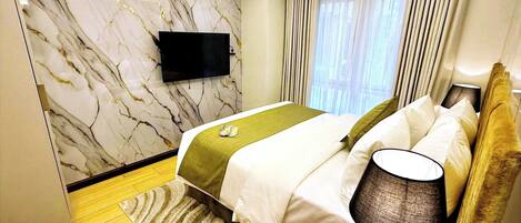 A hotel room with a marble wall, a bed with white bedding, a green throw, a nightstand with a lamp, and a flat-screen TV mounted on the wall.