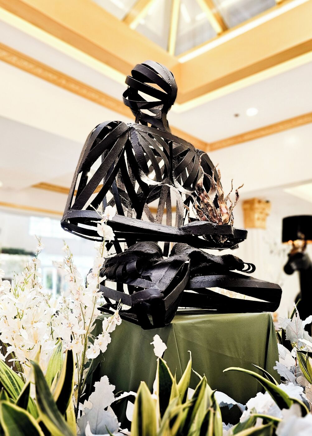 A sculpture of a human figure with a tray, surrounded by white flowers and greenery, in a well-lit indoor space.