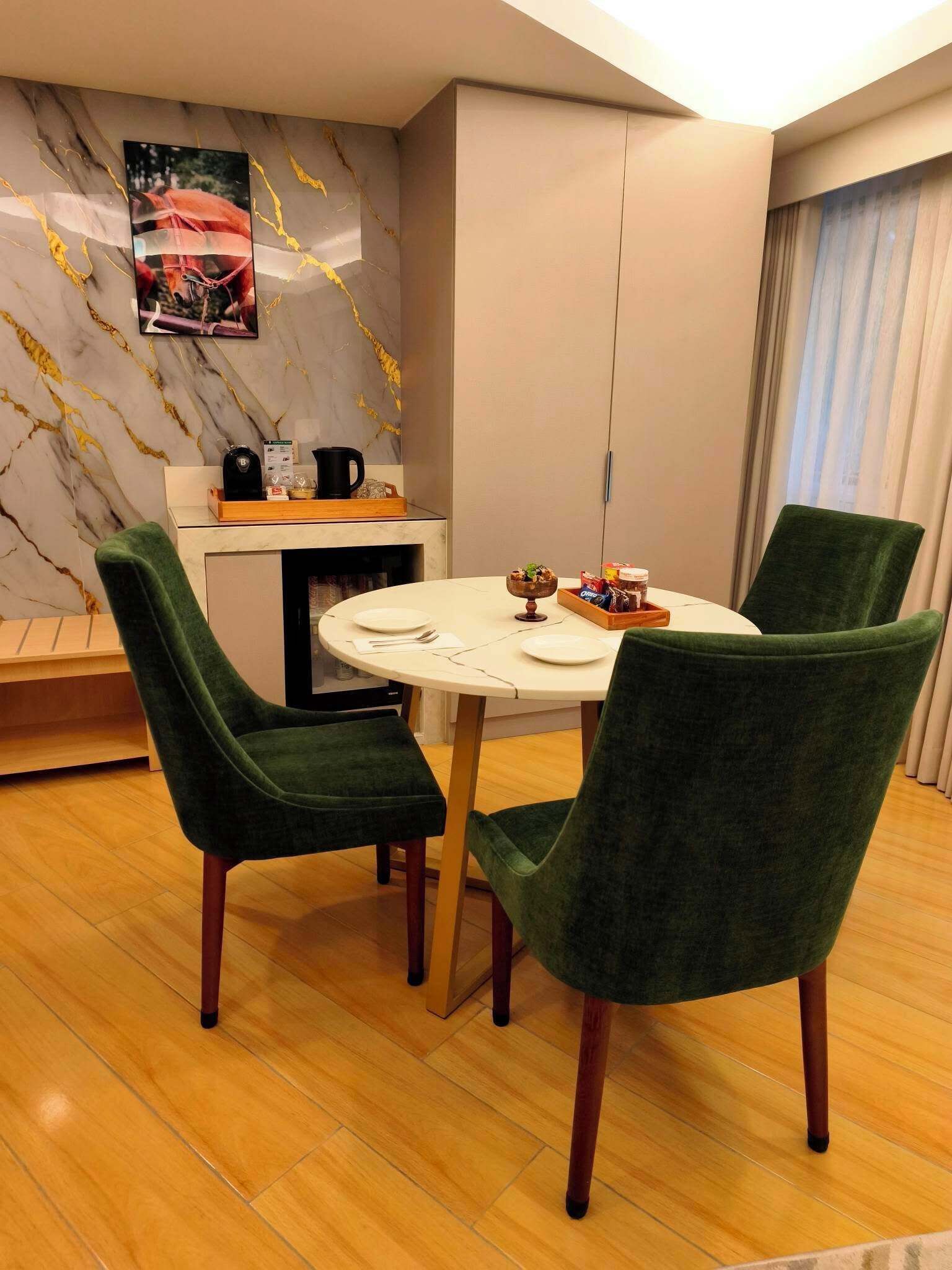 A dining area with a round table, four green upholstered chairs, a marble wall with a picture, and a kitchen area with a coffee machine.