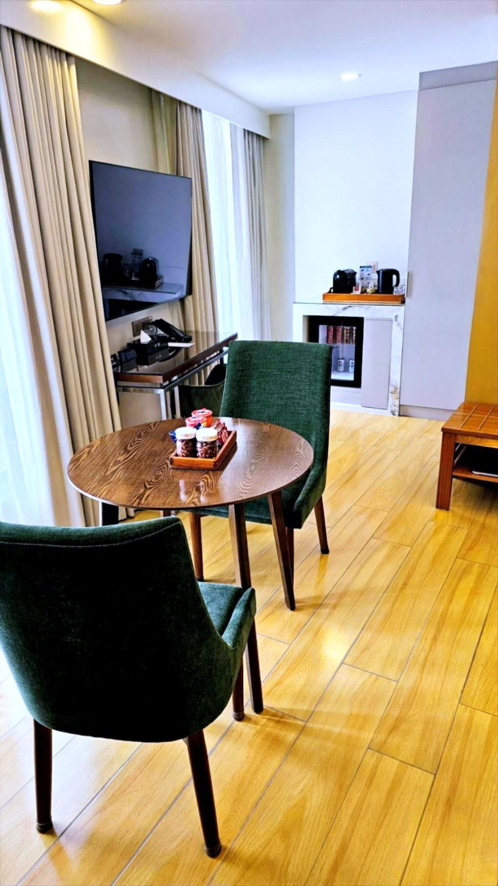 A modern hotel room with a wooden dining table, two green upholstered chairs, a flat-screen TV mounted on the wall, a small kitchen area with a coffee machine, and a bookshelf.