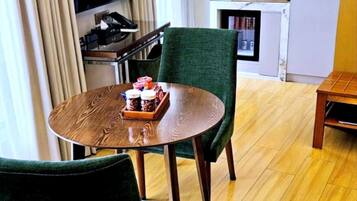 A modern hotel room with a wooden dining table, two green upholstered chairs, a flat-screen TV mounted on the wall, a small kitchen area with a coffee machine, and a bookshelf.