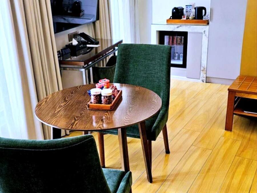A modern hotel room with a wooden dining table, two green upholstered chairs, a flat-screen TV mounted on the wall, a small kitchen area with a coffee machine, and a bookshelf.