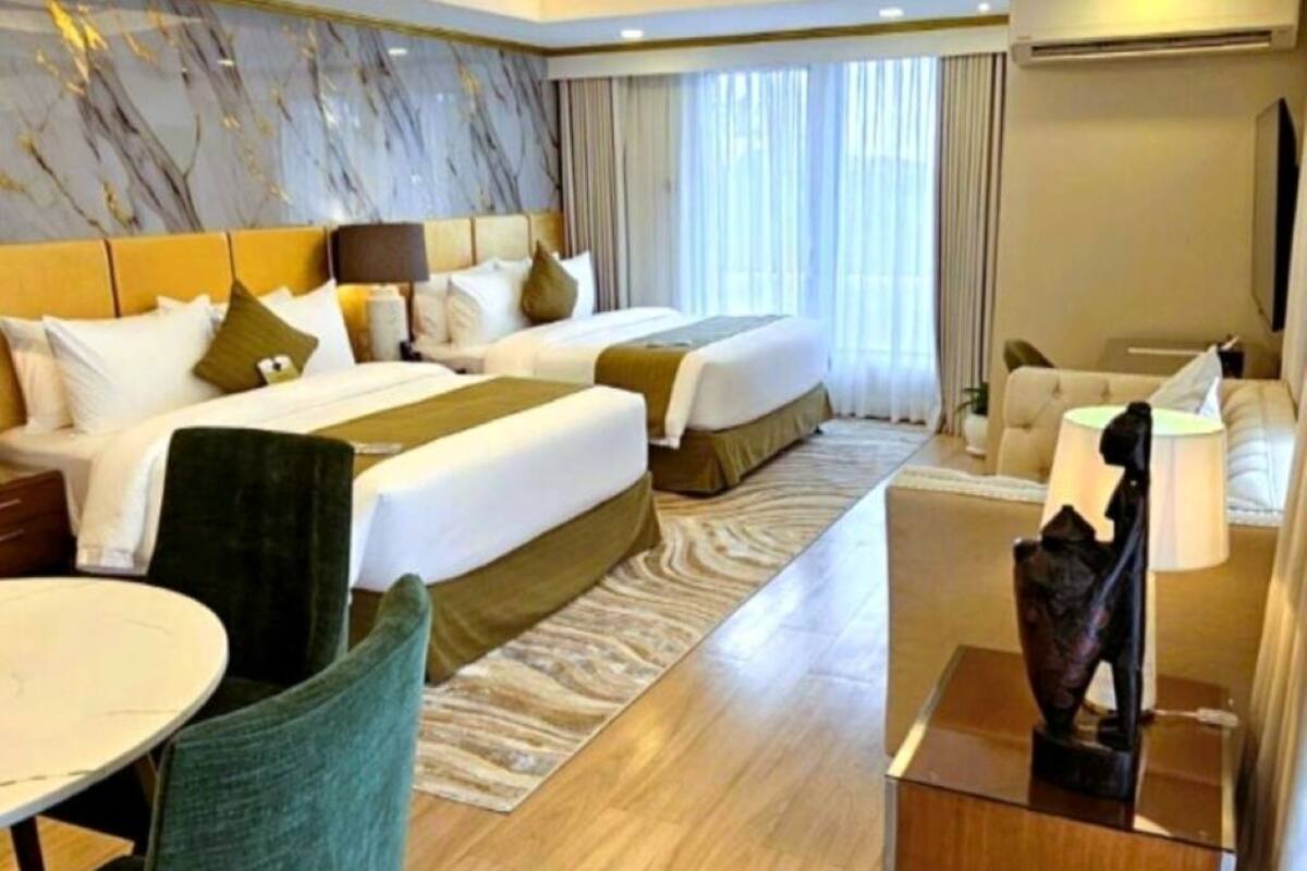 Ein Hotelzimmer mit zwei Betten, einer Sitzecke mit Tisch und Stühlen, einem Deckenventilator und einem großen Fenster mit Vorhängen.